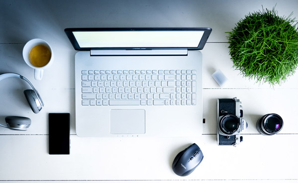 Skrivbord med laptop, hörlurar, mobiltelefon, kamera, kaffekopp och grön växt. Bilden är tagen uppifrån.