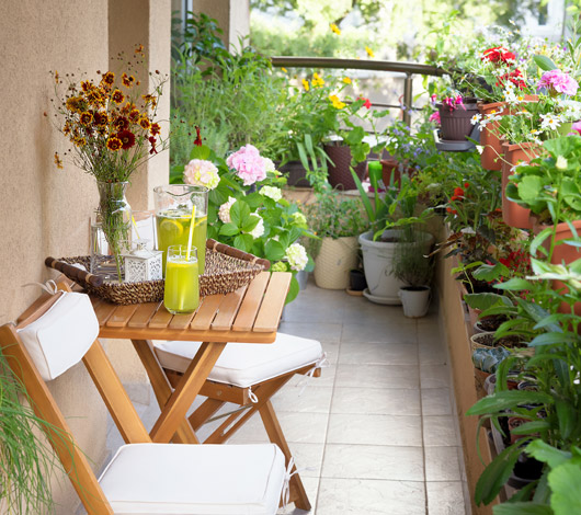 En grönskande balkong med träbord och stolar. På bordet en vas med ängsblommor och en saftbringare.