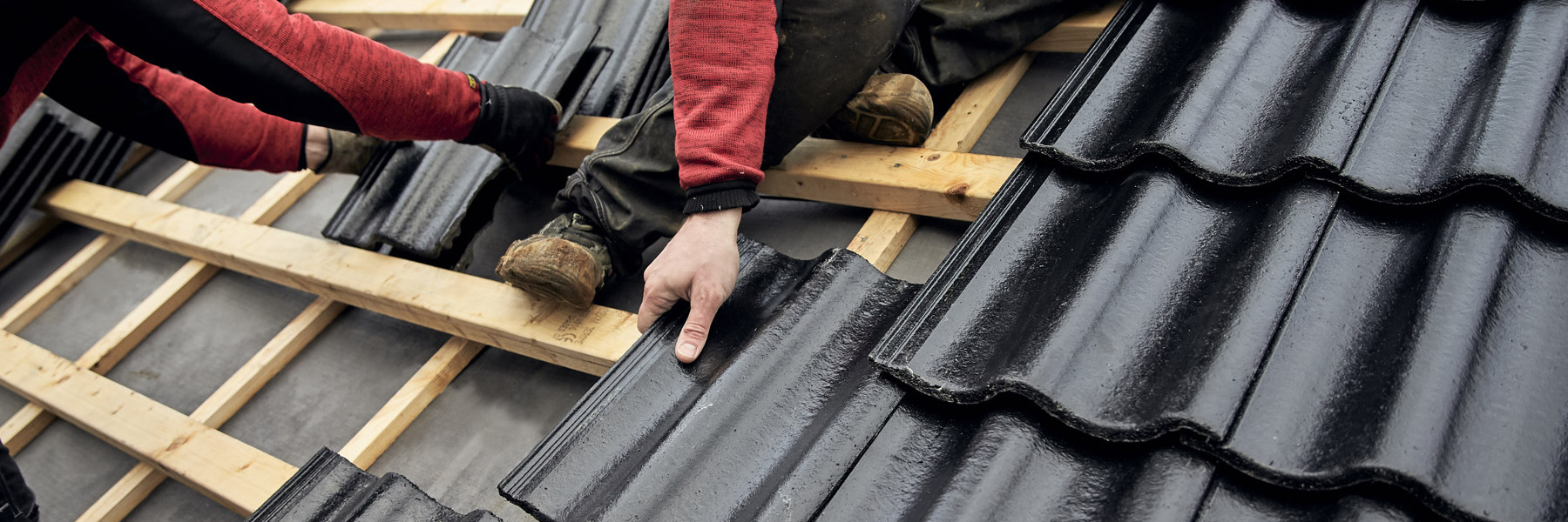 Närbild över takläggning, två män lägger svarta Aerlox betongpannor på läkten.