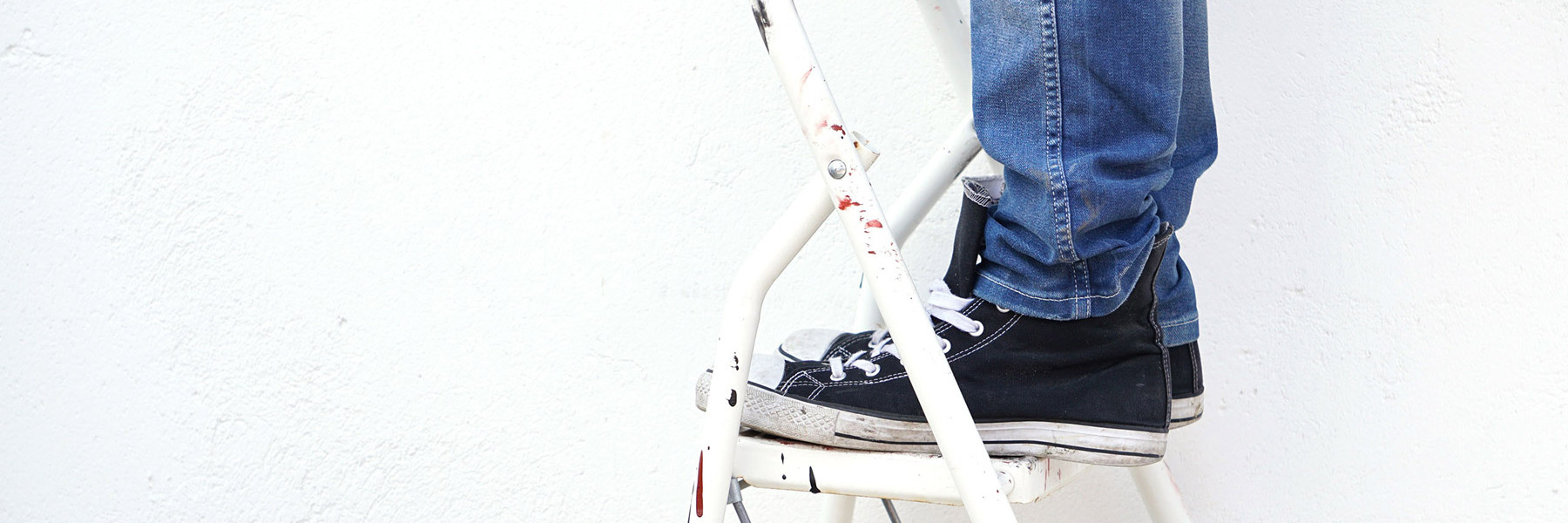 Någon med jeans och Converse-skor står på en stege. Man ser endast skor och byxbenen mot en vit vägg.